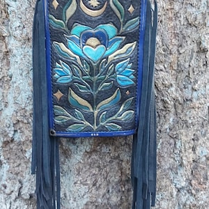 May include: A black leather crossbody bag with a blue floral design and fringe. The bag features a blue and gold floral pattern with a crescent moon and star design. The bag has a silver chain strap.