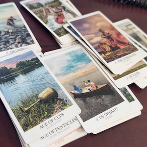 May include: A collection of tarot cards spread out, featuring various illustrations including the Ace of Cups, 6 of Swords, Strength, and 5 of Cups. Each card displays a unique scene with figures, landscapes, and water, with the card titles printed in black text at the bottom of each white-bordered card.