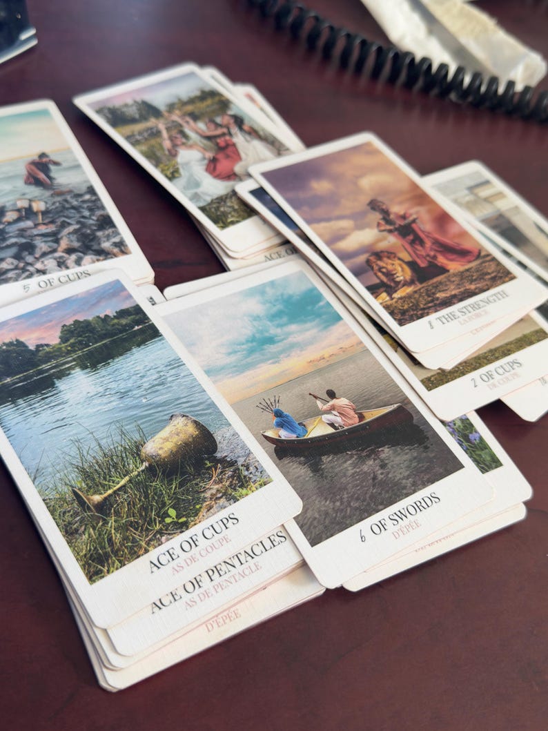 May include: A collection of tarot cards spread out, featuring various illustrations including the Ace of Cups, 6 of Swords, Strength, and 5 of Cups. Each card displays a unique scene with figures, landscapes, and water, with the card titles printed in black text at the bottom of each white-bordered card.