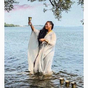 May include: A person wearing a white, flowing, sheer robe with gold trim, standing in water while holding a gold chalice aloft. Three additional gold chalices are positioned on a rock in the foreground. The text 4 OF CUPS DE COUPE is at the bottom.