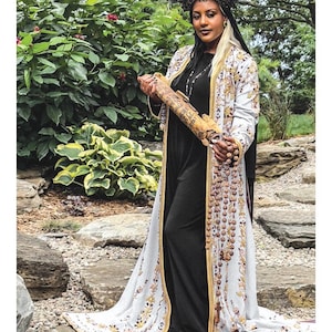 May include: A person wearing a long, white, embroidered coat over a black dress, holding a scroll. The person wears a dark, spiked crown. The text at the bottom reads 2 THE HIGH PRIESTESS LA PAPESSE. The attire features intricate gold and brown patterns.