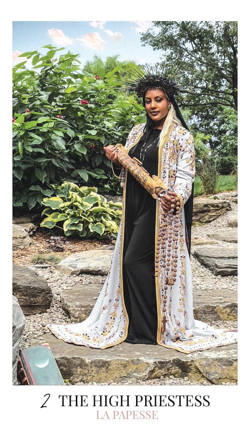 May include: A person wearing a long, white, embroidered coat over a black dress, holding a scroll. The person wears a dark, spiked crown. The text at the bottom reads 2 THE HIGH PRIESTESS LA PAPESSE. The attire features intricate gold and brown patterns.