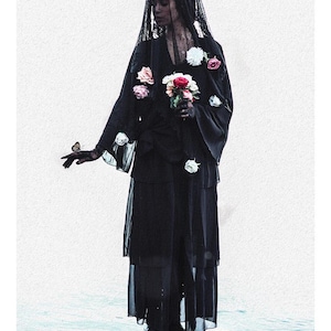 May include: A figure wearing a black layered dress, a black lace veil, and a crown, holding a bouquet of white and red roses. A small butterfly rests on one hand. The text 13 DEATH LA MORT is printed at the bottom of this tarot card design.