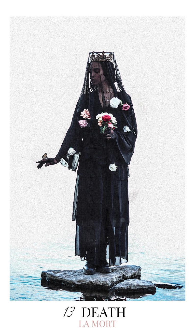 May include: A figure wearing a black layered dress, a black lace veil, and a crown, holding a bouquet of white and red roses. A small butterfly rests on one hand. The text 13 DEATH LA MORT is printed at the bottom of this tarot card design.
