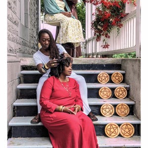May include: A tarot card titled 8 OF PENTACLES DE PENTACLE featuring four individuals on a porch. One person in a red dress sits on the stairs next to eight circular wooden discs, each engraved with a pentacle symbol. Others are seated nearby.