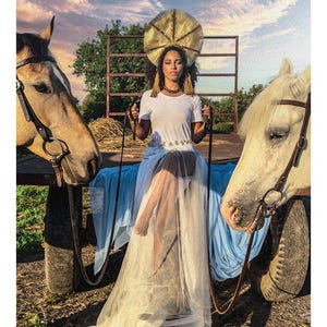 May include: A person wearing a white t-shirt and a long, flowing light blue tulle skirt with a gold halo-style headpiece, sitting on a cart between a tan horse and a white horse. The text 7 THE CHARIOT LE CHARIOT is printed at the bottom.