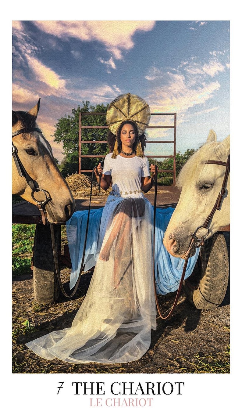May include: A person wearing a white t-shirt and a long, flowing light blue tulle skirt with a gold halo-style headpiece, sitting on a cart between a tan horse and a white horse. The text 7 THE CHARIOT LE CHARIOT is printed at the bottom.