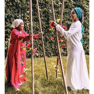 May include: A tarot card titled 4 OF WANDS DE B&Acirc;TON featuring two individuals standing outdoors, one in a red dress and head covering, the other in a white dress and teal head covering, holding four tall wooden poles adorned with red flowers.