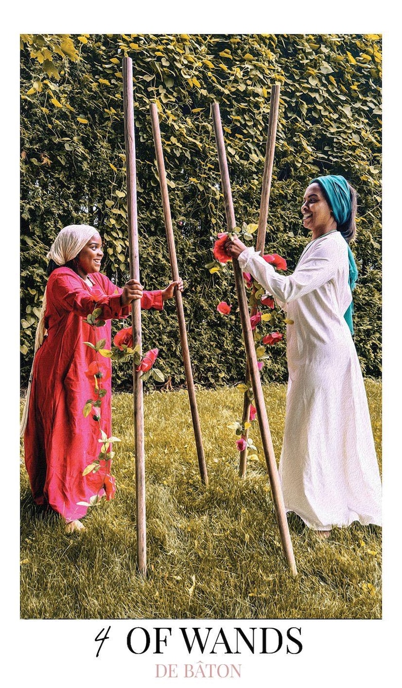 May include: A tarot card titled 4 OF WANDS DE B&Acirc;TON featuring two individuals standing outdoors, one in a red dress and head covering, the other in a white dress and teal head covering, holding four tall wooden poles adorned with red flowers.