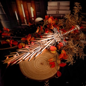 Op de afbeelding: Een gedetailleerd, sierlijk zwaard met een zilveren lemmet en een decoratieve greep. Het zwaard ligt op een houten schijf, omringd door herfstbladeren met rode en oranje bladeren. Op de achtergrond zijn boeken en donkere stof te zien.
