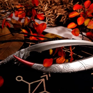 May include: A silver and red decorative dagger with ornate detailing, resting on a patterned black surface. The curved blade is paired with a matching sheath, accented with red and white designs. Autumn leaves and branches surround the dagger.