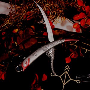 May include: A decorative, silver-colored knife with a curved blade and ornate handle. The blade is white, and the handle has red accents and engraved designs. The knife is displayed against a dark background with red leaves and foliage.