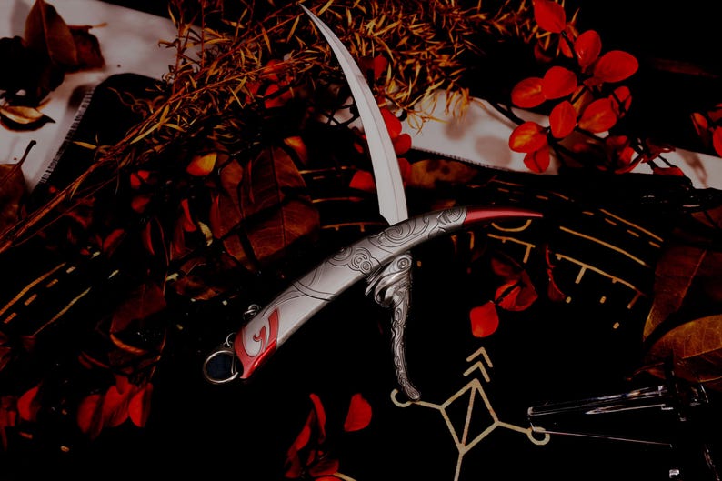 May include: A decorative, silver-colored knife with a curved blade and ornate handle. The blade is white, and the handle has red accents and engraved designs. The knife is displayed against a dark background with red leaves and foliage.