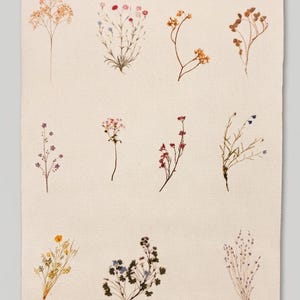 May include: A rectangular off-white fabric wall hanging featuring a grid of eleven distinct pressed wildflower illustrations. The botanical prints include various stems, buds, and small blossoms in shades of yellow, pink, purple, and brown.