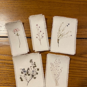 May include: Five small rectangular fabric tags with frayed edges, each featuring a different pressed flower design. The flowers are arranged in a natural, organic style, with delicate stems and petals.