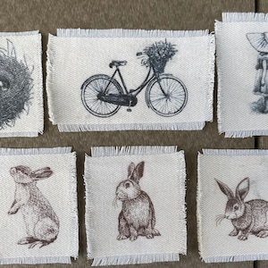May include: Six fabric squares with different designs. The first square shows a bird's nest with eggs. The second square shows a bicycle with a basket of flowers. The third square shows a girl in a dress and hat. The fourth square shows a brown rabbit standing on its hind legs. The fifth square shows a brown rabbit sitting. The sixth square shows a brown rabbit sitting with its ears up.