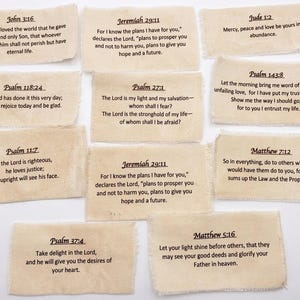 May include: A collection of ten rectangular beige fabric labels with frayed edges, each printed with black text featuring various Bible verses including John 3:16, Jeremiah 29:11, Jude 1:2, Psalm 118:24, Psalm 27:1, Psalm 143:8, Psalm 11:7, Matthew 7:12, Psalm 37:4, and Matthew 5:16.