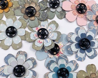 以下が含まれることがあります： 中央に黒い金属製のスナップボタンが付いた、重ねられた紙の花の装飾品のコレクション。花には、水玉模様や植物のプリントなど、青、緑、ピンク、黄色の色合いを含むさまざまなパターンと色が表示されています。
