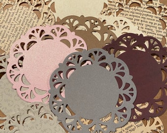 May include: A collection of decorative paper doilies in various colors, including cream, brown, pink, gray, and burgundy. The doilies have intricate, cut-out floral patterns and are arranged on a background of vintage book pages.
