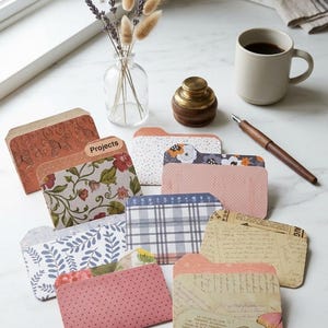 May include: A collection of small, rectangular paper file folders featuring various patterns including floral, plaid, polka dot, and vintage script designs. One folder tab is clearly labeled with the word Projects in black sans-serif text.