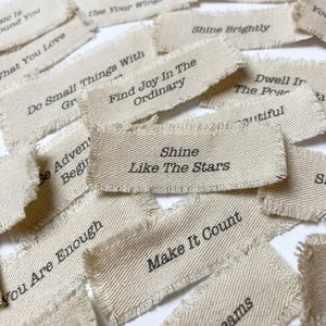 May include: A collection of rectangular, off-white fabric labels with frayed edges, each featuring black printed text including phrases like Shine Like The Stars, Find Joy In The Ordinary, Make It Count, Shine Brightly, and You Are Enough.