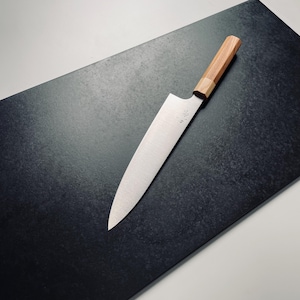 May include: A silver chef's knife with a wooden handle rests on a black cutting board. The knife has a slightly curved blade and a pointed tip.