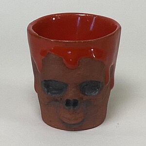 May include: A small, red ceramic shot glass shaped like a skull with dripping red glaze. The skull has black eye sockets and a small, open mouth.