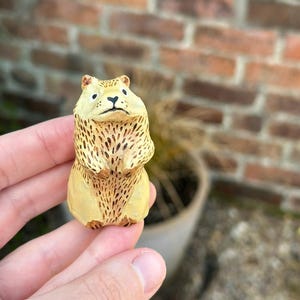 Könnte beinhalten: Eine kleine, handgeschnitzte Holzfigur eines braunen und gelben Bären mit schwarzen Augen. Der Bär steht aufrecht mit seinen Pfoten vor sich.