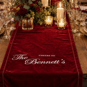 May include: A deep red velvet table runner featuring the white embroidered text "CHEERS TO The Bennett's" centred on a rustic wooden dining table. The table is set with gold-rimmed plates, dark red napkins, crystal glassware, and floral centrepieces.