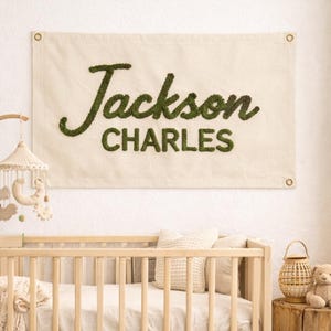 May include: A nursery scene featuring a light wood crib with bedding, a mobile, and a banner that reads "Jackson CHARLES" in green. A small teddy bear sits on a wooden stump next to the crib. The room has a neutral color palette.