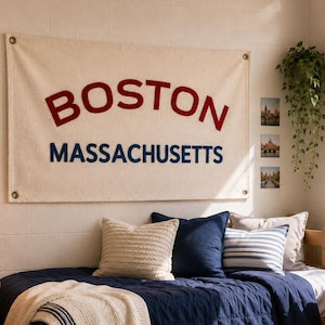 Op de afbeelding: Een rechthoekige wandbanner van gebroken witte stof met het woord BOSTON in grote rode letters en MASSACHUSETTS in kleinere blauwe letters, voorzien van metalen ringen in de hoeken, opgehangen boven een bed met marineblauw beddengoed.