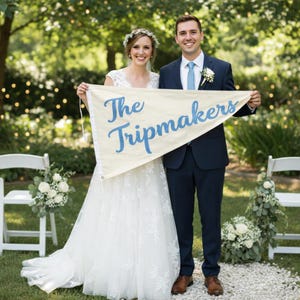 Op de afbeelding: Een stel houdt een spandoek vast met de tekst "The Tripmakers" in blauwe letters. De vrouw draagt een witte trouwjurk en een bloemenkrans. De man draagt een marineblauw pak met een lichtblauwe stropdas. Witte stoelen en bloemstukken zijn op de achtergrond.