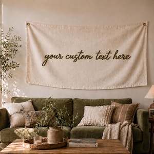 May include: A rectangular beige fabric wall hanging featuring the dark green text your custom text here in a cursive script font. The banner is suspended by two small metal rings at the top corners against a plain light-coloured wall.