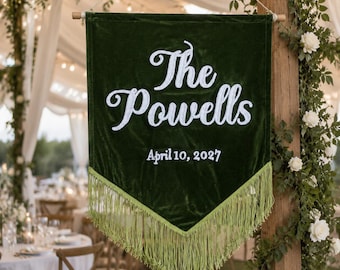 May include: A dark green velvet pennant banner featuring white embroidered text that reads The Powells and the date April 10, 2027. The banner has a pointed bottom edge finished with long, light green fringe and hangs from a wooden dowel rod.
