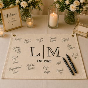 May include: A cream-colored wedding guest book with the initials "L|M" and the year "EST 2025". The book is surrounded by pens, candles, and floral arrangements. The words "Welcome to our Wedding" are on a framed sign.