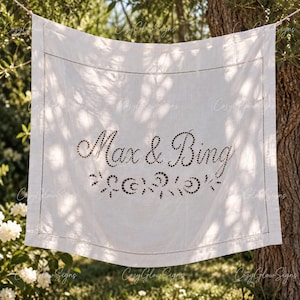 Può includere: Un cartello in tessuto bianco di forma quadrata con la scritta Max & Bing in un carattere corsivo scuro e punteggiato. Il design include un motivo floreale punteggiato decorativo sotto i nomi e un bordo cucito lungo il perimetro del tessuto.