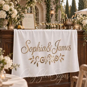May include: A white fabric banner featuring the gold text Sophia & James in a cursive font, accented with a gold mosaic-style floral design below the names. The banner is draped over a dark wooden bar counter in an outdoor setting.