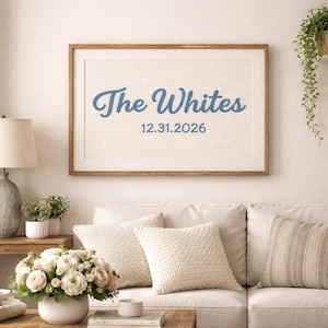 Puede incluir: Una obra de arte enmarcada con el texto "The Whites" en azul y la fecha "12.31.2026". La obra se exhibe sobre un sofá blanco con cojines decorativos. También se ven una lámpara, plantas y un arreglo floral.