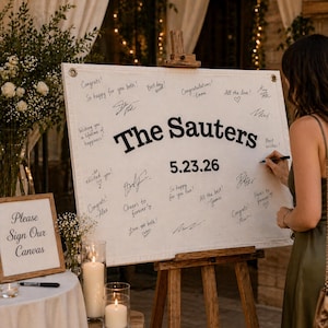 Puede incluir: Un letrero de boda de lona blanca sobre un caballete de madera, firmado por los invitados. El letrero dice "The Sauters" y "5.23.26" en letras negras. El texto adicional incluye "Please Sign Our Canvas".
