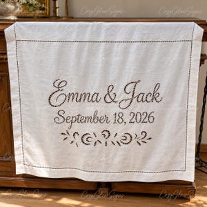 May include: A white rectangular linen runner featuring dark brown embroidered text that reads Emma & Jack and September 18, 2026. The fabric includes a decorative cutwork floral motif below the date and a stitched border frame.