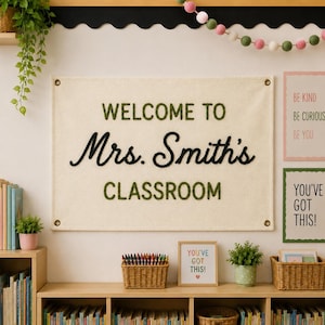 Puede incluir: Un estandarte de pared rectangular de tela color hueso con el texto bordado en verde "WELCOME TO" y "CLASSROOM" y el texto negro en cursiva "Mrs. Smith's" en el centro. El estandarte tiene ojales metálicos en las cuatro esquinas.