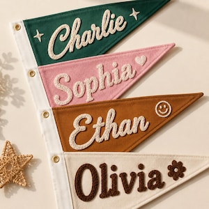 Peut inclure: Quatre fanions triangulaires en feutrine avec des œillets blancs sur une bande verticale. Le haut est vert foncé avec le texte blanc Charlie et des étoiles. En dessous, rose avec le texte blanc Sophia et un cœur. Puis marron avec le texte blanc Ethan et un smiley. En bas, crème avec le texte marron Olivia et une fleur.