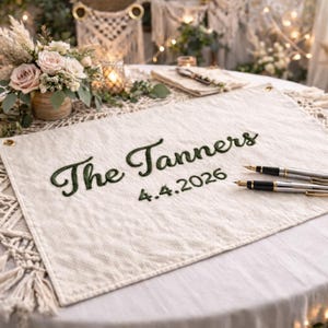 Peut inclure: Livre d'or de mariage crème avec l'inscription brodée "The Tanners 4.4.2026" en vert. Deux stylos à plume reposent sur le livre. La table est décorée de fleurs, de bougies et de macramé.