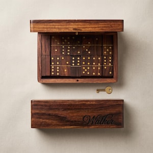 May include: A dark brown wooden domino set housed in a rectangular box with a hinged lid. The dominoes feature gold-coloured pips. A small gold-coloured metal key is included. The box lid is engraved with the name Walker in a cursive script font.