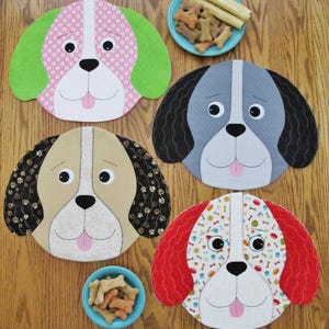May include: Four dog-shaped placemats with different patterns. The placemats are made of fabric and have a stitched design. The placemats are on a wooden table with two bowls of dog biscuits.