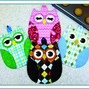 May include: Four colorful owl-shaped pot holders. The owls have large eyes and are made of patterned fabric in green, blue, pink, and brown. A baking sheet with cookies is in the background. Each pot holder has a loop for hanging.