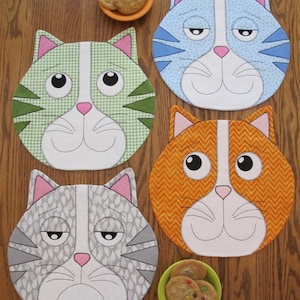 Może przedstawiać: Cztery okrągłe podkładki pod talerze w kształcie kota w różnych kolorach i wzorach. Podkładki są wykonane z tkaniny i mają haftowane detale. Podkładki są ułożone w kółko na drewnianej powierzchni.