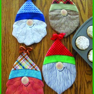 Puede incluir: Cuatro agarraderas de olla acolchadas con forma de gnomos con sombreros y barbas de diferentes colores. Las agarraderas están sobre una superficie de madera.