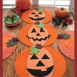 Puede incluir: Tres manteles individuales de color naranja en forma de calabaza con caras de calabaza sonriente negras. Los manteles individuales están sobre una mesa de madera con una pequeña calabaza naranja y flores rojas.