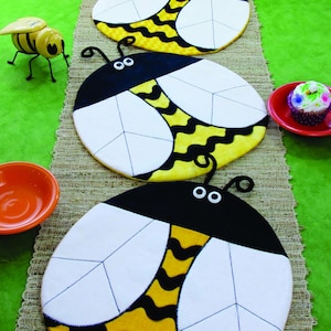May include: Three yellow and black bee-shaped placemats with white wings and black eyes. The placemats are on a brown woven surface.